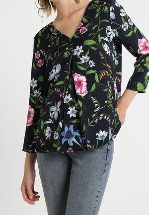 Kvinde iført en sort bluse med lange ærmer og farverige blomsterprint samt lysegrå jeans, stående med den ene hånd i lommen.