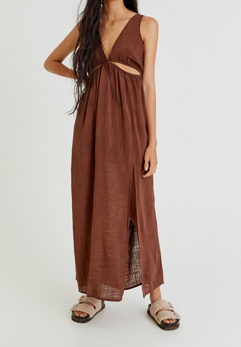 Robe maxi marron avec un décolleté en V profond et un détail découpé, fabriquée en tissu léger. Comprend une fente latérale et une surface texturée.