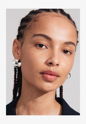 Silver hoop earrings with a textured, sculptural design. Features a shiny finish and smooth curves, contrasting with braided hair.