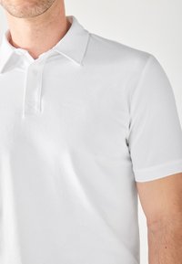 Camisa polo blanca de algodón con cuello clásico, mangas cortas, tapeta de botones y una textura suave. Presenta dobladillos de doble costura.