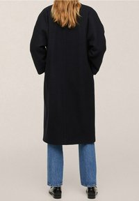 Long manteau noir à coupe décontractée, avec des épaules tombantes et une silhouette droite. Associé à un jean bleu clair et des chaussures noires.