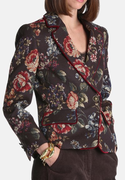 Femme portant un blazer croisé à motif floral foncé avec passepoil rouge et boutons dorés, assorti à un pantalon foncé et un bracelet doré épais au poignet gauche.
