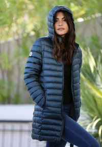 Manteau rembourré bleu marine avec capuche ; design matelassé, texture brillante et poches latérales. Porté sur un haut noir et associé à un jean bleu.