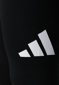 Juodos leggingai, pagaminti iš tempimo audinio, su dideliu baltu trijų juostų logotipu, esančiu dešinės kojos šone.