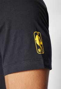 Camiseta de manga corta negra con un parche amarillo del logo de la NBA en la manga izquierda, hecha de una tela suave y lisa con un corte clásico.