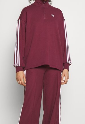 Bordowy dres Adidas z białymi paskami na rękawach i nogawkach, z pulowerem na suwak sięgający do ćwiartki oraz szerokimi spodniami.