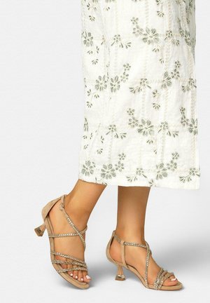 Voeten in beige sandalen met hak en bandjes, gecombineerd met een witte rok tot aan de enkel met geborduurde bloemmotieven.