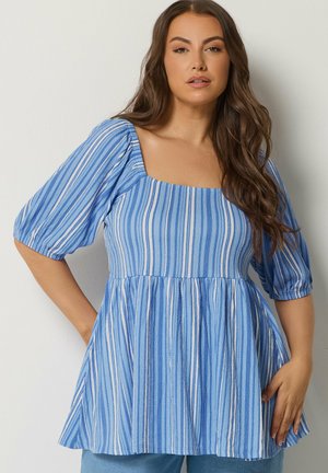 Femme aux longs cheveux bruns portant un haut à rayures verticales bleues et blanches avec des manches bouffantes et un décolleté carré, debout contre un mur clair.