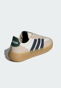 Světlé béžové tenisky s námořnicky modrými pruhy, bílými stranami a zeleným akcentem na patě. Mají gumovou podrážku a texturované semišové prvky.