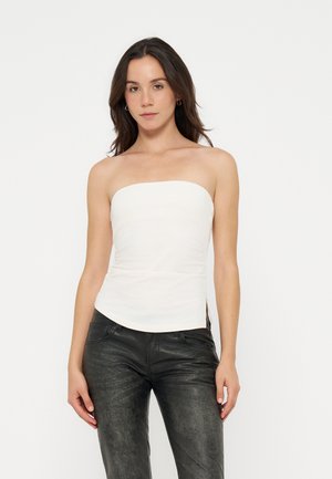 Femme aux cheveux foncés portant un top blanc sans bretelles et un pantalon noir texturé, debout devant un fond clair uni.