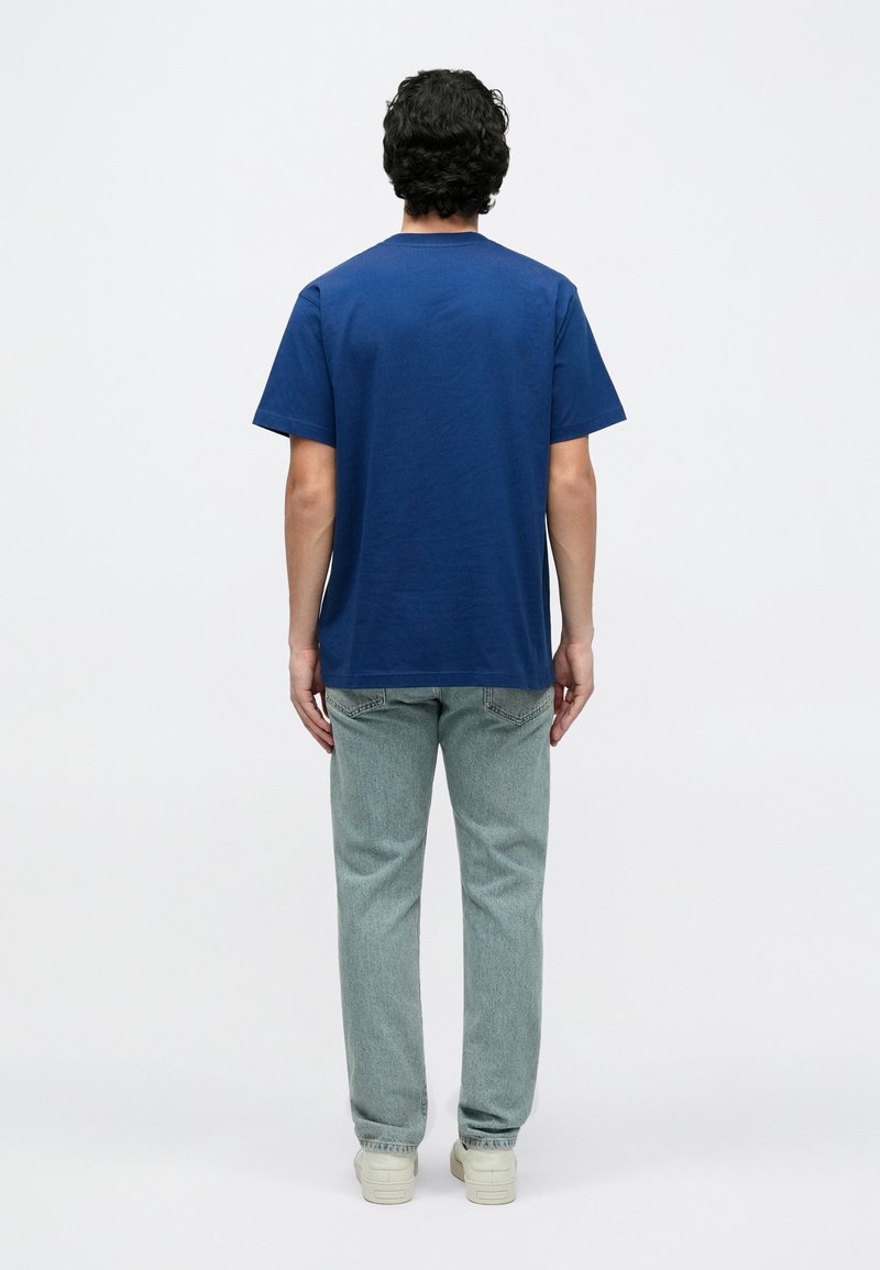 Blauw katoenen T-shirt met korte mouwen, ronde hals en een losse pasvorm. Draagt lichtblauwe spijkerbroek en witte sneakers.