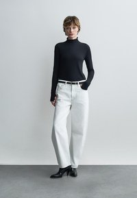 Noor seisab ühevärvilise seina taustal, kandes musta kõrge kaelusega kampsunit, valgeid laiade säärtega teksapükse, musta vööd ja musti terava ninaga saapaid.