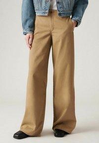 Wijduitlopende broeken in beige katoen, met een hoge taille, voorzakken en een strakke afwerking. Gecombineerd met een spijkerjack en zwarte schoenen.