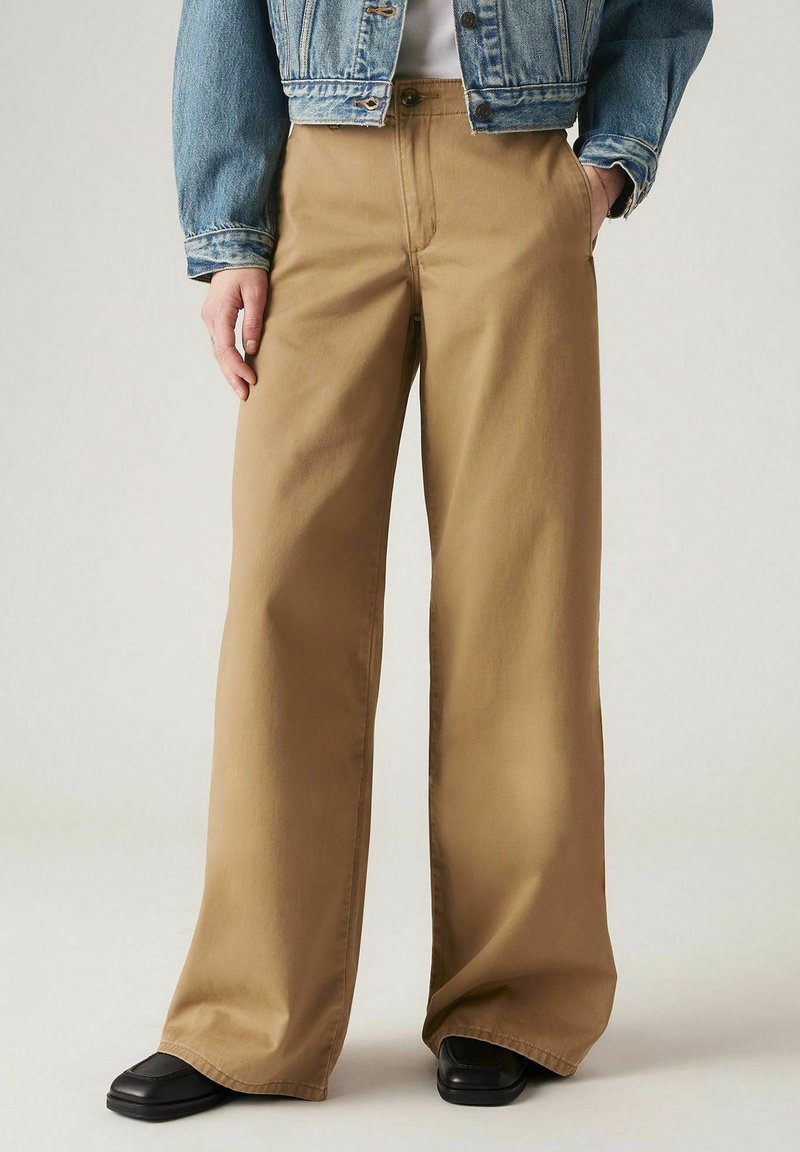 Wijduitlopende broeken in beige katoen, met een hoge taille, voorzakken en een strakke afwerking. Gecombineerd met een spijkerjack en zwarte schoenen.