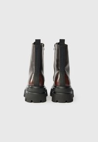 Sötétbarna bőr bokacsizma, vastag fekete talppal és hátsó cipzárral, fehér háttér előtt.