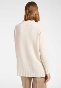 Frau mit schulterlangem braunem Haar, die einen cremefarbenen gerippten Rollkragenpullover und hellbraune Hosen trägt, von hinten gesehen.