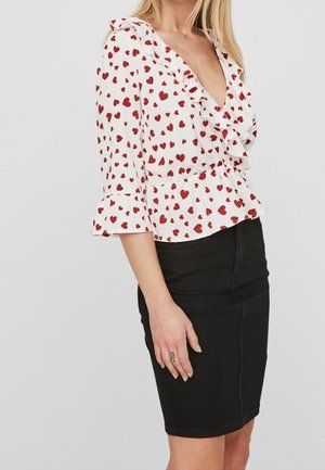 Femme portant un chemisier blanc à motifs de cœurs rouges et à volants, associé à une jupe noire longueur genou, debout devant un fond uni.