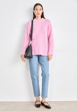 Rosa flauschiger Pullover mit hohem Kragen, hellblaue Jeans und schwarze Ballerinas. Der Pullover hat eine lockere Passform und gerippte Bündchen. Schwarze Handtasche mit Tragegurt.