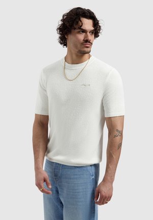 Man met krullend haar die een witte, textuurrijke korte mouw trui draagt, lichtblauwe spijkerbroek, gouden ketting en gouden ringen, met tatoeages op zijn arm.