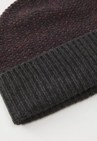Knitted beanie in dark gray with textured pattern featuring small red and purple flecks. Ribbed cuff adds detail and structure.