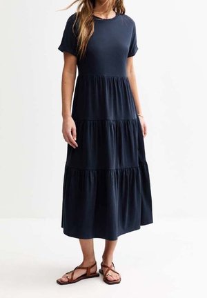 Frau in einem navyblauen, gestuften Midikleid mit kurzen Ärmeln und braunen, flachen Sandalen, die vor einem schlichten weißen Hintergrund steht.
