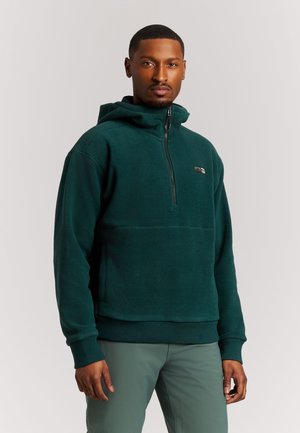 Man met een donkergroene fleece hoodie met halve rits en groene broek, staand tegen een effen achtergrond