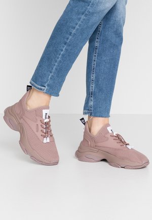 Osoba nosząca różowe masywne sneakersy z fakturowaną siateczką i niebieskie dżinsy, stojąca na tle jednolitego jasnego tła.