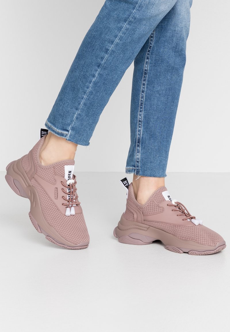Personne portant des baskets roses épaisses avec un mesh texturé et un jean bleu, debout devant un fond clair uni.