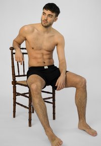 Černé bavlněné kraťasy s látkovým páskem, které nosí sedící mužský model na dřevěné židli na jednobarevném pozadí.