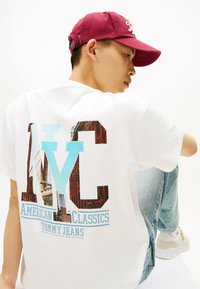 Witte katoenen t-shirt met een grafische "NYC" print met fotografische elementen. Gecombineerd met lichtblauwe jeans en een bordeauxrode pet.