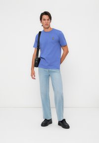 T-shirt en coton bleu à manches courtes avec un logo jaune sur la poitrine, associé à un jean droit bleu clair et des chaussures noires.