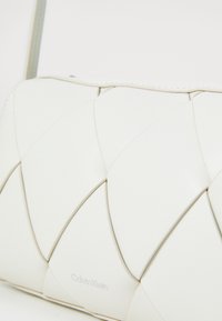 Borsa a clutch in pelle intrecciata bianca con il sottile logo Calvin Klein davanti, su uno sfondo chiaro e semplice.