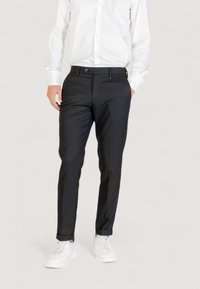 Pantalones negros ajustados con una textura suave, corte slim, bolsillos frontales y cierre de botón, combinados con zapatillas blancas y una camisa blanca.