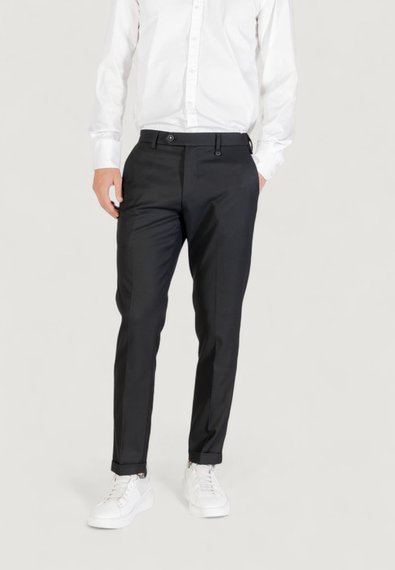 Pantalones negros ajustados con una textura suave, corte slim, bolsillos frontales y cierre de botón, combinados con zapatillas blancas y una camisa blanca.
