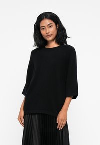 Pull noir oversize en tricot doux, avec un col rond et des manches trois-quarts. Texture lisse et coupe décontractée.