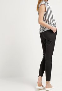 Femme portant un pantalon noir slim, des chaussures blanches sans lacets, et un haut sans manches rayé noir et blanc, debout les mains dans les poches.