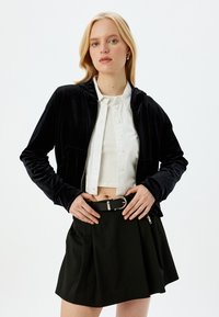 Giacca cropped in velluto nero con cappuccio, abbinata a una camicia bianca con bottoni e una gonna nera. Caratterizzata da una zip e tasche laterali.