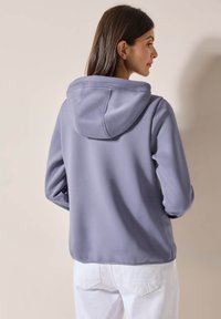 Felpa viola lavanda con coulisse al collo, texture morbida e maniche lunghe. L'orlo è leggermente curvato sul retro.