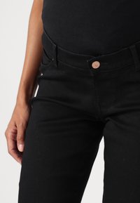 Jeans de mezclilla negra con un diseño ajustado, que cuentan con un cierre de botón, bolsillos cosidos y una textura suave. Mano descansando sobre el muslo.