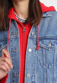 Jeansjacka med silverknappar, bröstfickor, matchad med en röd zip-up sweatshirt med vit krage och röda snörningar.