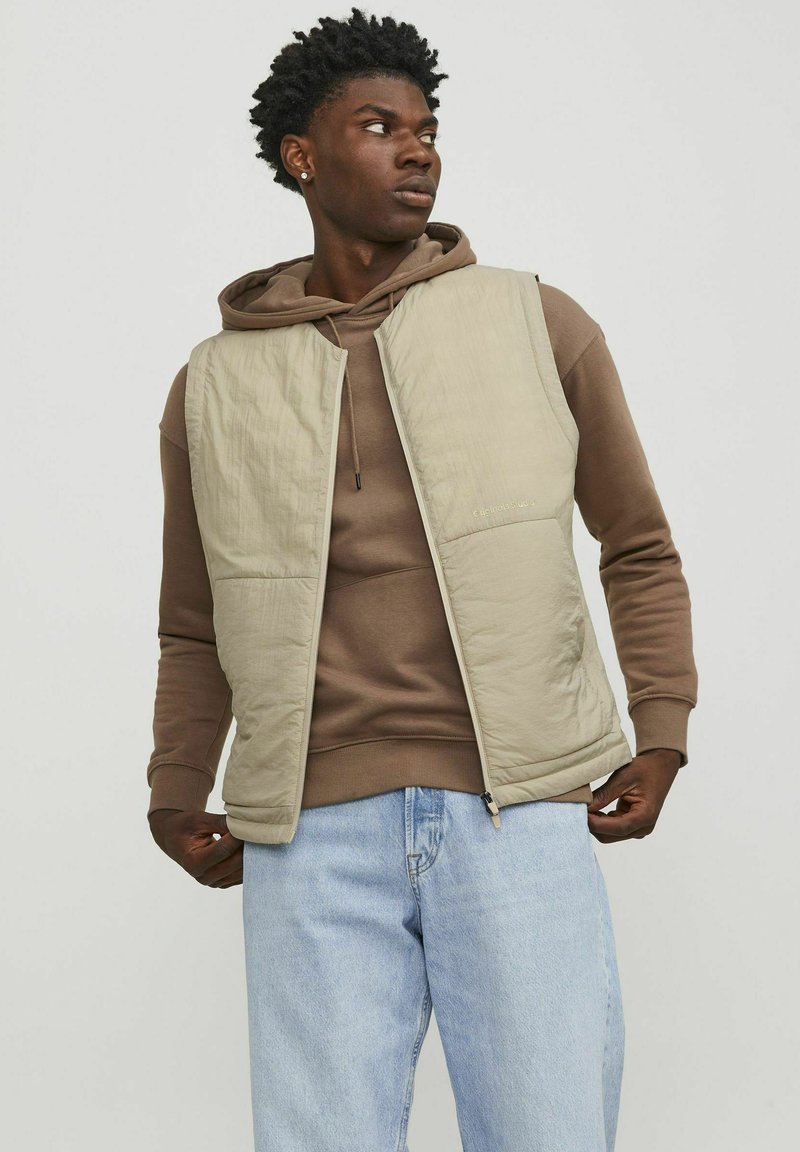 Gilet matelassé beige sur un hoodie marron, avec fermeture éclair et poches latérales. Porté avec un jean bleu clair. Matière texturée et légère.