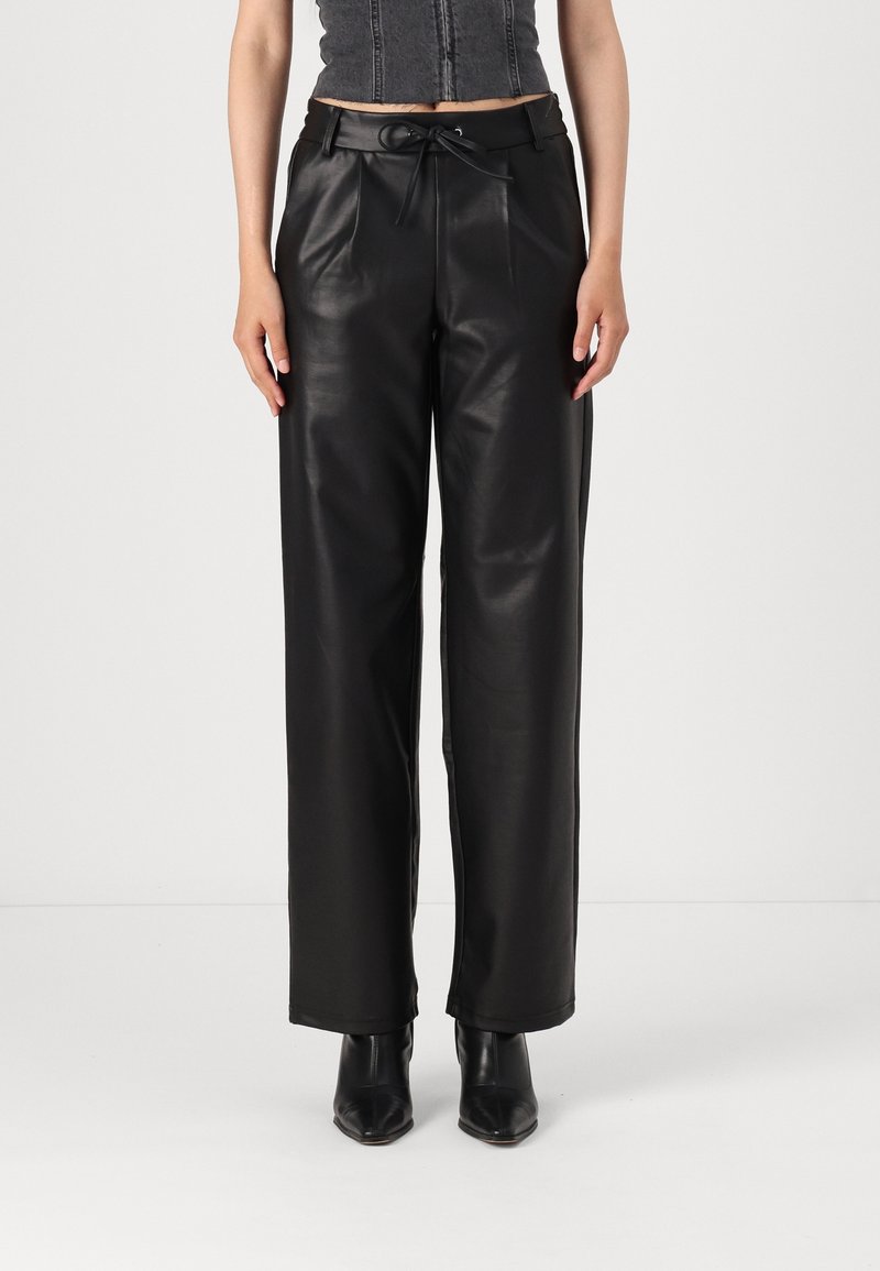 Femme portant un pantalon en similicuir noir taille haute avec une ceinture nouée, associé à des bottines noires à bout pointu et un top court.