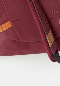 Marondfarvet stoftaske med brune læder detaljer. Har en glat tekstur, buet design og kontrasterende syninger. Inkluderer justerbar strop.