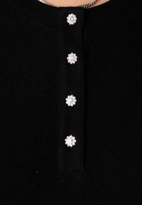 Zwart wollen kledingstuk met vier decoratieve zilveren bloemvormige knopen en een zichtbare zilveren kettinghalsband aan de bovenkant.