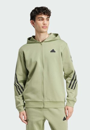 Młody mężczyzna ubrany w jasnooliwianą bluzę Adidas z kapturem oraz pasujące spodnie z czarnymi paskami na rękawach i logo Adidas.