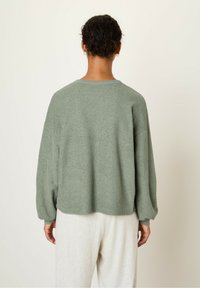Maglione verde, texture morbida con una vestibilità rilassata, spalle scese e scollatura arrotondata, abbinato a pantaloni della tuta grigi chiari.