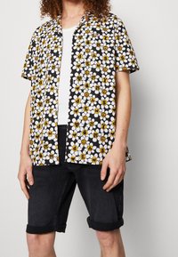 Koszulka z krótkim rękawem na czarnym tle, w białe i żółte wzory kwiatowe, z literami i symbolami, zestawiona z ciemnymi szortami denimowymi.