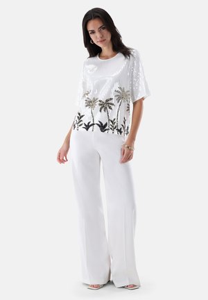 Frau trägt ein weißes Pailletten-Top mit Palmenmotiven und weiten weißen Hosen, steht vor einem schlichten hellen Hintergrund.