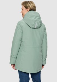 Persoon in een lichtgroene hoodie met reflecterende details op de capuchon, gezien vanaf de achterkant tegen een effen achtergrond.