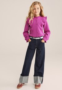 Vijolična srajca s teksturo in nabrano obleko na ramenih, v kombinaciji s temnimi širokimi hlačami, ki imajo svetel jeans rob in rjave superge.